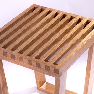 Könnte beinhalten: Ein handgefertigter Holzschemel mit Lattenrost und quadratischen Beinen. Das hellbraune Holz hat eine natürliche Maserung und ist mit sichtbaren Schwalbenschwanzverbindungen verbunden. Der Hocker ist etwa 46 cm hoch.