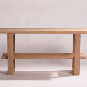 Op de afbeelding: Een lichtgekleurde houten console met een rechthoekig blad en een onderste plank. De tafel heeft vier vierkante poten en een eenvoudig, minimalistisch ontwerp. De plank heeft een lattenontwerp. De tafel is ongeveer 122 cm lang.