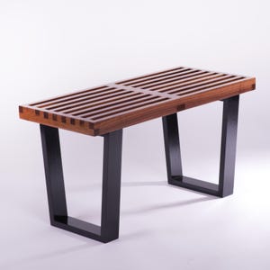 Peut inclure: Un banc en bois avec un dessus à lattes et des pieds noirs. Le banc est en bois brun foncé avec un design rectangulaire. Les pieds sont de couleur noire contrastante, créant une esthétique moderne. Le banc mesure environ 122 cm de long.