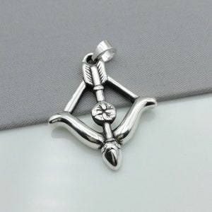 May include: A silver-colored pendant featuring a bow and arrow design. The arrow is centered within the bow, with a detailed fletching. The pendant has a loop at the top for a chain. The background is a solid gray.
