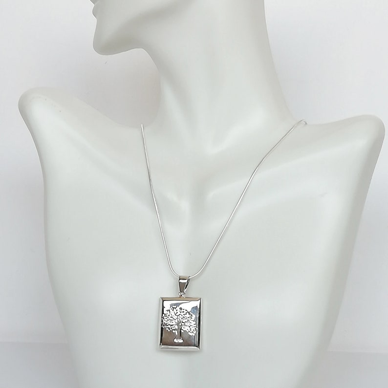 May include: A silver necklace with a square pendant featuring a detailed tree design.