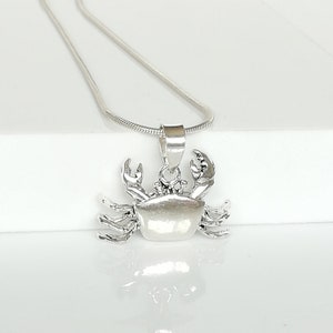 May include: A silver-colored crab pendant necklace. The crab pendant is detailed with claws and a textured body. The necklace chain is a thin, silver snake chain. The pendant is displayed on a white surface.