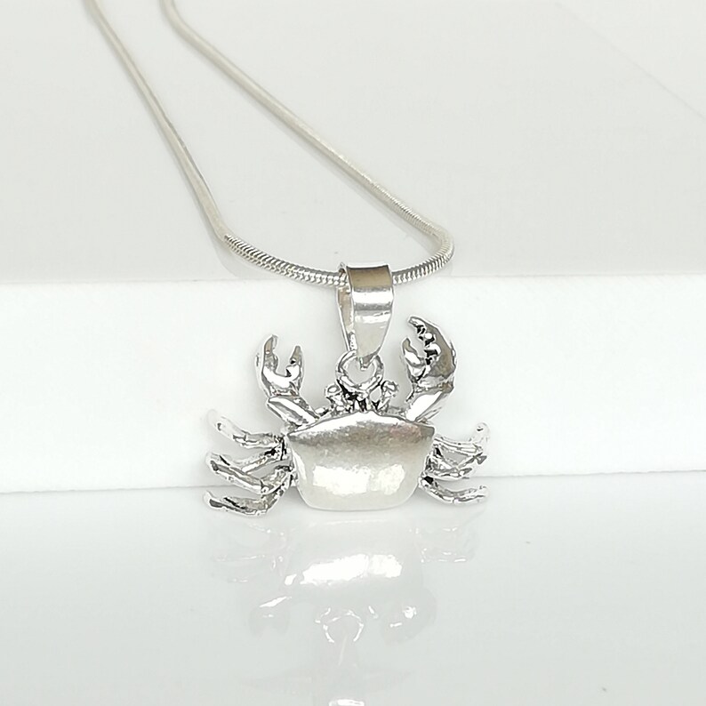 May include: A silver-colored crab pendant necklace. The crab pendant is detailed with claws and a textured body. The necklace chain is a thin, silver snake chain. The pendant is displayed on a white surface.