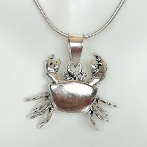 May include: A silver-colored crab pendant with intricate detailing, suspended from a thin, silver chain. The crab's claws are raised, and its body is textured. The pendant is likely made of sterling silver or a similar metal. The necklace is displayed against a white background.