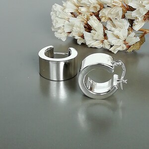Könnte beinhalten: Ein Paar silberne Creolen mit einem dicken, breiten Band. Die Ohrringe sind aus glänzendem Metall und haben ein schlichtes, modernes Design.