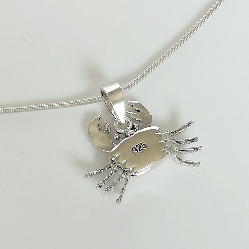May include: A silver crab pendant necklace. The pendant is detailed with claws, legs, and a textured body. The back of the pendant is stamped with "925". The necklace chain is a thin, silver snake chain.