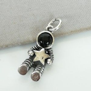 Puede incluir: Un charm de astronauta plateado con un casco de piedra negra y una estrella dorada en el pecho. El charm tiene un diseño detallado y acanalado, y un pequeño lazo para colgar.