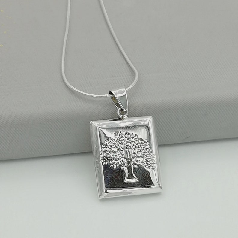 May include: A silver necklace with a rectangular pendant featuring a detailed tree design. The pendant is attached to a silver chain.