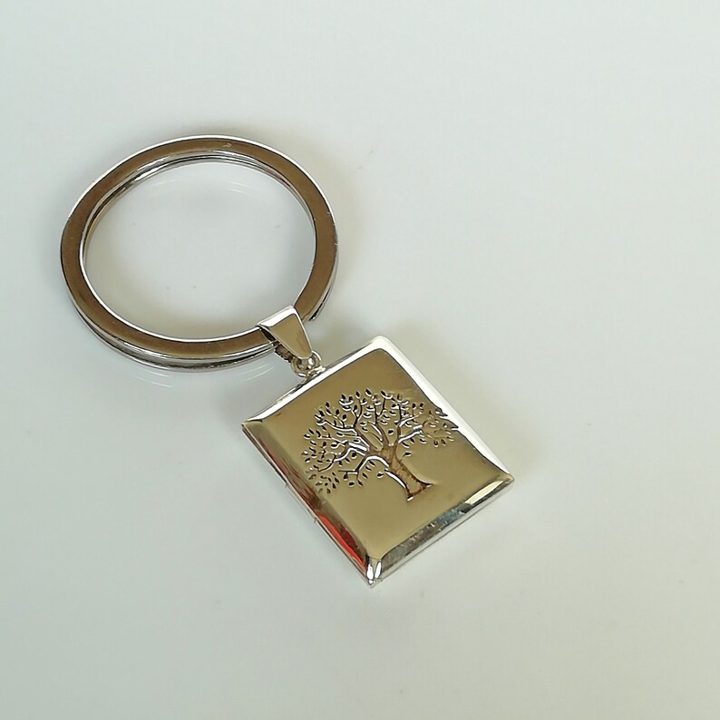 May include: Silver keychain with a tree of life design. The keychain has a silver ring and a rectangular charm with an engraved tree.