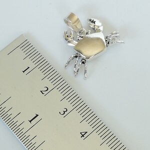 May include: A silver crab pendant with a gold-colored body, shown next to a ruler. The pendant is approximately 1 inch in length. The crab has detailed claws and legs, and a loop for a chain. The background is a light, neutral color.