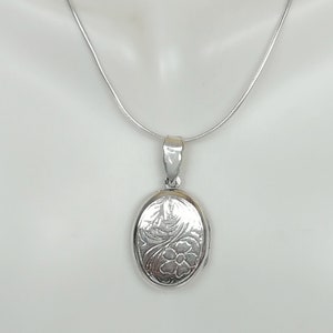 Puede incluir: Un collar de cadena de plata con un colgante de medallón ovalado de plata. El medallón tiene un diseño floral grabado en la parte delantera.