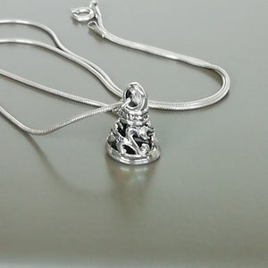May include: A silver pendant necklace featuring a bell-shaped charm with intricate openwork designs. The pendant hangs from a delicate, snake chain. The necklace is displayed on a neutral, reflective surface.