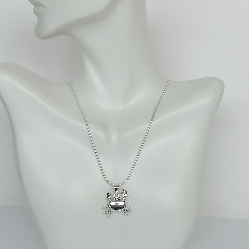 May include: A silver-colored crab pendant necklace. The crab pendant has detailed claws and a small gemstone. The necklace is displayed on a white mannequin. The chain is a thin, silver snake chain.