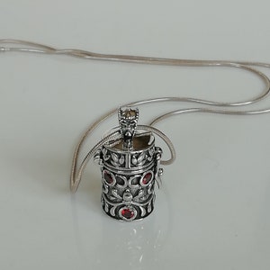 Op de afbeelding: Zilveren ketting met een gedetailleerde hanger met een kleine, cilindrische doos met rode edelstenen. De doos heeft een ingewikkeld ontwerp en lijkt een medaillon of een bedel te zijn.