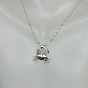 May include: A silver crab pendant necklace. The pendant is a detailed crab design with claws and legs. The necklace chain is a thin, silver snake chain. The pendant is approximately 1 inch wide.