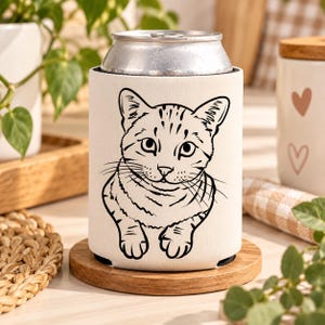 以下が含まれることがあります： 前面にタビーキャットの黒い線画イラストが描かれた白いフォーム製の缶クーラーカバー。このカバーは円筒形で、標準的な金属製の飲料缶にフィットするように設計されており、飲み物を保温・保冷するためのものです。