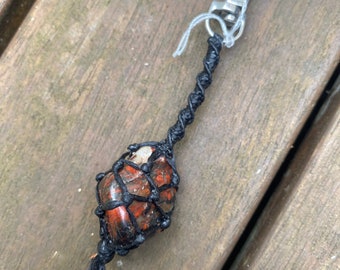 May include: A red, black, and white stone wrapped in black cord with a silver key chain. The stone is tied with a knot at the top and bottom. The keychain is labeled "crafted".