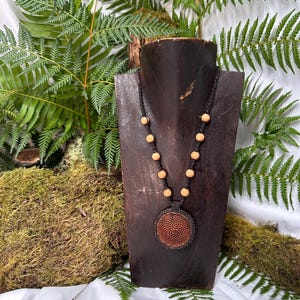 Op de afbeelding: Een handgemaakte ketting met een grote, ronde hanger met een geweven, roodbruin ontwerp. De ketting heeft een zwart koord met lichtgekleurde houten kralen. De hanger wordt getoond op een donkere houten buste tegen een achtergrond van groene varens.