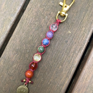 Puede incluir: Un llavero de cuentas con una serie de cuentas de piedras preciosas de colores en una fila, conectadas por un cordón rojo. Las cuentas incluyen piedras claras, moradas, azules, verdes, amarillas, naranjas y rojas. Un cierre dorado y un dije de carita sonriente completan el diseño.