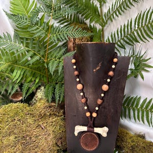 May include: A handcrafted necklace featuring a bone pendant and a circular, woven element. The necklace has a brown cord with wooden and off-white beads. The pendant is displayed on a dark brown wooden stand, with a backdrop of ferns and moss.
