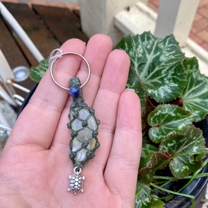 Puede incluir: Un llavero de macramé verde con un cristal transparente y un dije de tortuga plateado. El llavero está unido a un anillo de llave plateado.