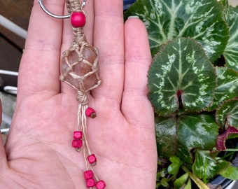 May include: A tan macrame keychain with a pink stone and pink beads. The keychain is held in a hand.