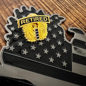 May include: A black and silver metal pin featuring a US flag design and the word "RETIRED" on a gold ribbon. The pin is adorned with a gold eagle and laurel wreath.