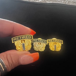 May include: Three gold-coloured military retirement pins with the word "RETIRED" in black lettering. Each pin features a stylised eagle with wings spread and a single black stripe.