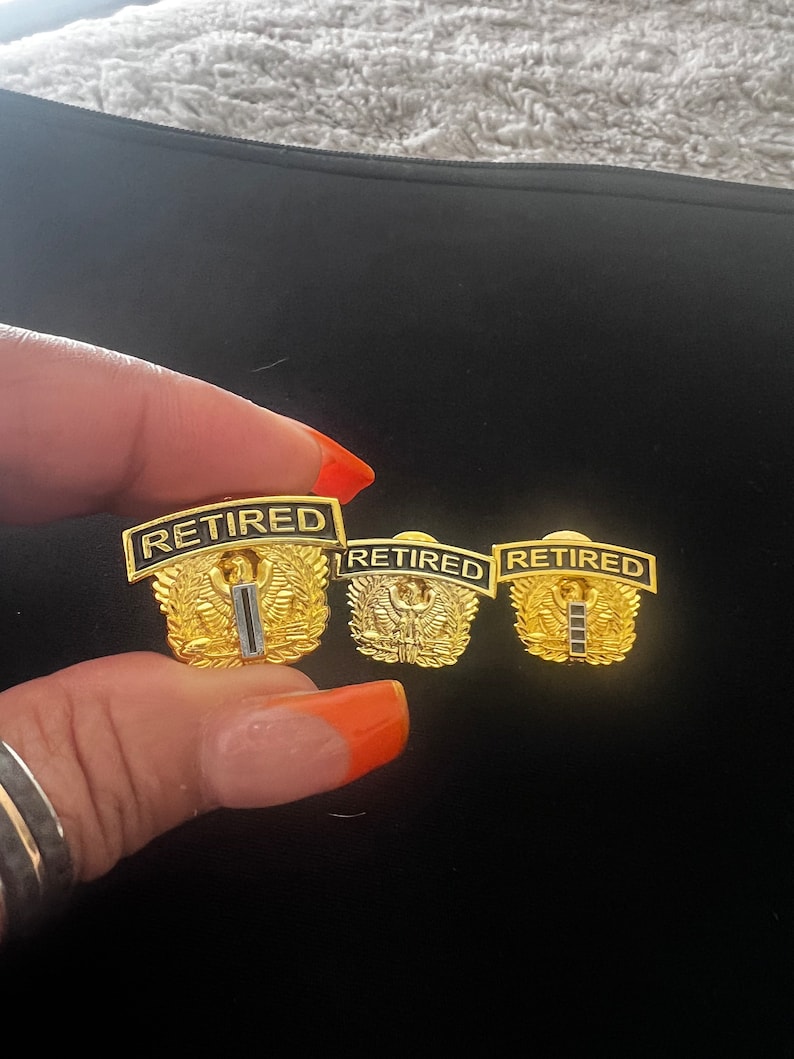 May include: Three gold-coloured military retirement pins with the word "RETIRED" in black lettering. Each pin features a stylised eagle with wings spread and a single black stripe.