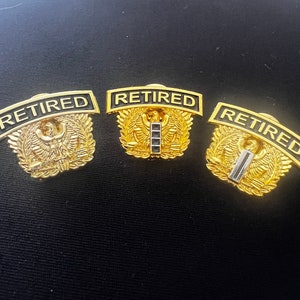 May include: Three gold-coloured military retirement pins with black lettering that reads "RETIRED". Each pin features a stylised eagle with wings outstretched and a laurel wreath.