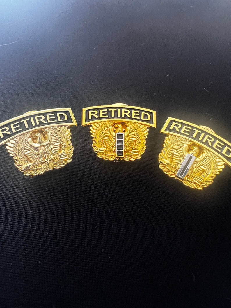 May include: Three gold-coloured military retirement pins with black lettering that reads "RETIRED". Each pin features a stylised eagle with wings outstretched and a laurel wreath.