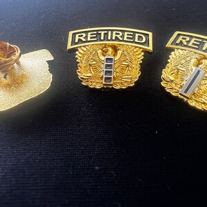 May include: Two gold-coloured military pins with the word "RETIRED" on a black banner. The pins feature a stylised eagle with wings spread and a gold bar with black stripes.
