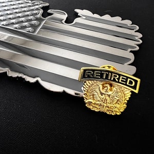 May include: A gold-coloured eagle pin with a black and white ribbon that says "Retired". The pin is on top of a silver-coloured American flag.