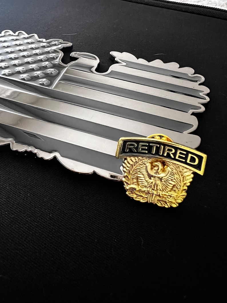 May include: A gold-coloured eagle pin with a black and white ribbon that says "Retired". The pin is on top of a silver-coloured American flag.