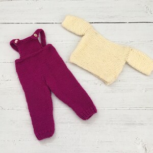 May include: A knitted baby outfit set with a pink knitted jumpsuit and a cream knitted top.