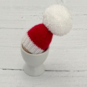 May include: A white ceramic egg cup with a red and white knitted Santa hat on top. The hat has a white pom-pom.