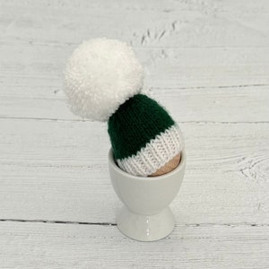 May include: A green and white knitted hat with a white pom pom on top, sitting in a white egg cup.