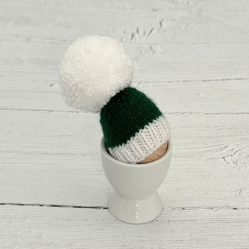 May include: A green and white knitted hat with a white pom pom on top, sitting in a white egg cup.