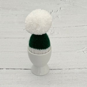 May include: A white ceramic egg cup with a green and white knitted hat with a white pom-pom on top.