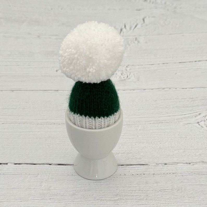 May include: A white ceramic egg cup with a green and white knitted hat with a white pom-pom on top.