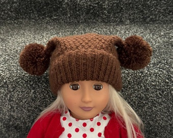 May include: A brown knitted hat with two pom-poms, worn by a doll. The hat is a dark brown colour and has a textured knit pattern. The doll is wearing a red cardigan over a white top with red polka dots.