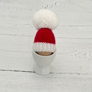 May include: A red and white knitted Santa hat with a white pom-pom sits in a white egg cup.