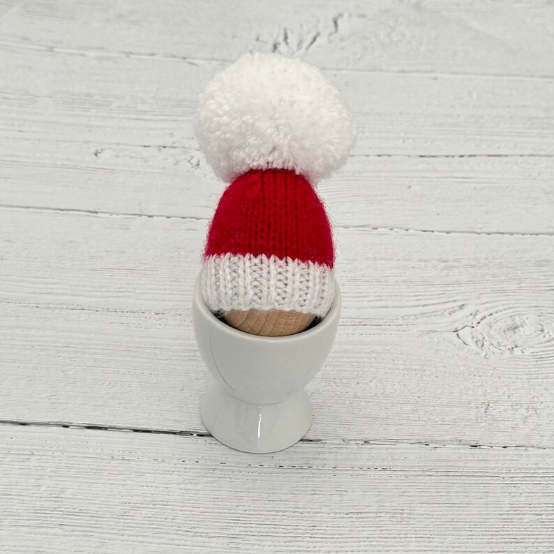 May include: A red and white knitted Santa hat with a white pom-pom sits in a white egg cup.
