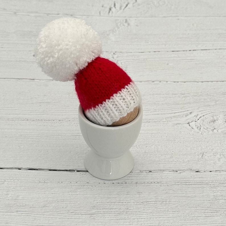 May include: A white egg cup with a red and white knitted Santa hat on top.