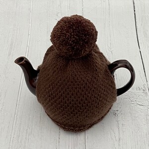 Puede incluir: Un calienta-tetera de punto marrón con un gran pompón en la parte superior.