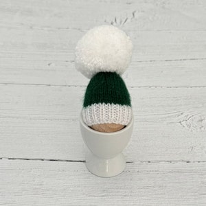 May include: A green and white knitted hat with a white pom pom on top, sitting in a white egg cup.