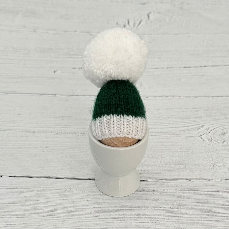 May include: A green and white knitted hat with a white pom pom on top, sitting in a white egg cup.