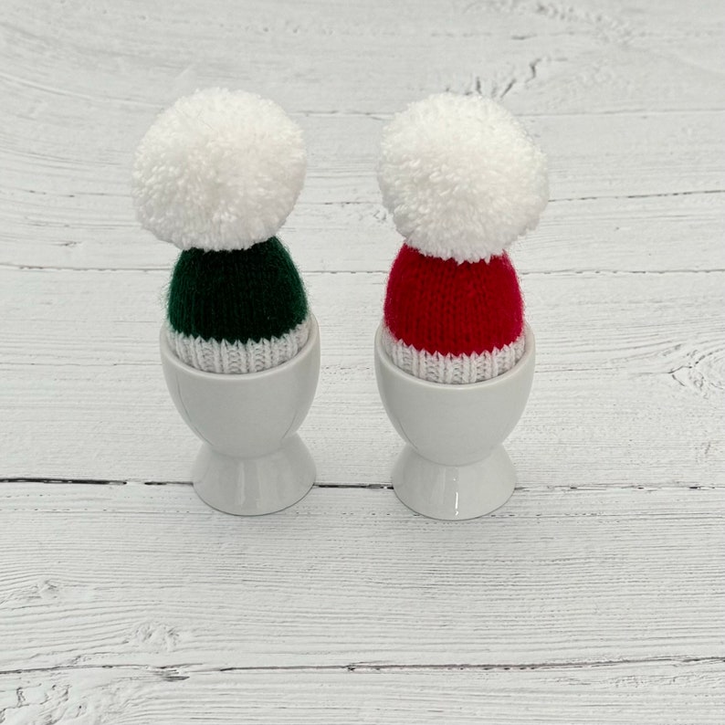 May include: Two white ceramic egg cups with knitted hats. One hat is green with a white pom-pom and the other is red with a white pom-pom.