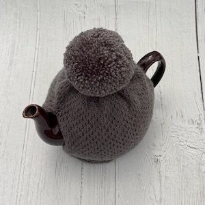 May include: A grey knitted tea cosy with a large pom-pom on top, covering a brown ceramic teapot. The teapot has a curved handle and a spout. The cosy is designed to keep tea warm.