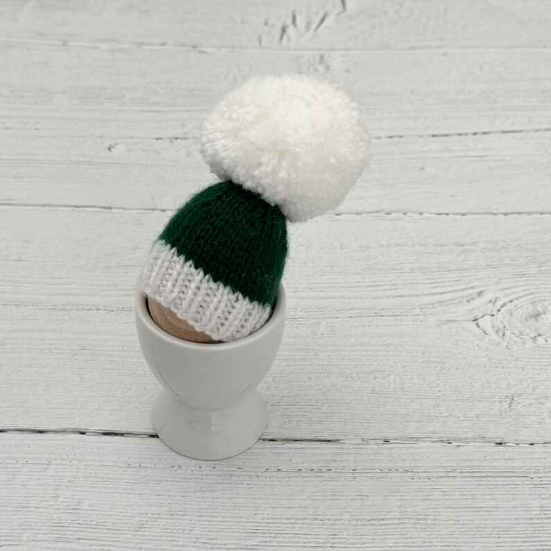 May include: A green and white knitted hat with a white pom pom sits in a white egg cup on a white wooden surface.